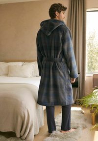 Kariertes Bademantel in dunklem Blau und Grautönen, mit Kapuze, Bindegurt und langen Ärmeln, kombiniert mit dunklen Hosen, auf einem Teppich neben einem Bett.