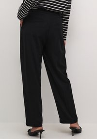 Pantalons larges noirs en tissu lisse, dotés de poches arrières. Le modèle a associé des talons noirs à enfiler et un haut rayé.