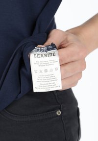 Marinblå T-shirt med skötselrådsetikett där "SEASIDE"-logotypen syns. Etiketten visar tygkomposition och skötselanvisningar. Svarta byxor syns i bakgrunden.