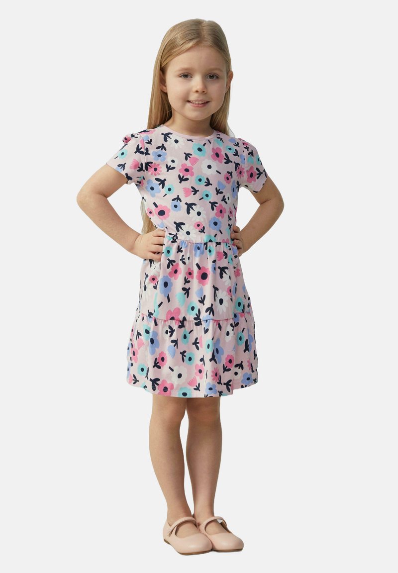 Blumenkleid aus leichtem Stoff, mit rosa, blauen und schwarzen Mustern, kurzen Ärmeln und einem schwingenden Rock. Die Schuhe sind beige Ballerinas.