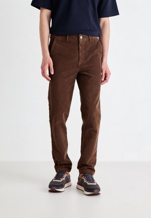 Homme portant un pantalon en velours côtelé marron, un t-shirt bleu marine et des baskets multicolores, debout devant un fond blanc.
