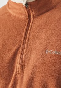 Pomarańczowy polar o odcieniu rdzy z kołnierzem na pół zamek, z kremowym wykończeniem kołnierza i srebrnym logo na piersi. Miękka tekstura.