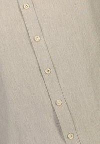 Camisa de color beige claro con tejido texturizado, que presenta una fila vertical de seis botones color crema y un patrón de tejido sutil.