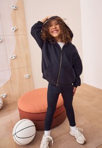 Sweat à capuche noir avec fermeture éclair et poches avant, porté avec des leggings bleu marine. Comprend un siège rond et rembourré orange ainsi qu'un ballon de basket blanc posé sur le sol.