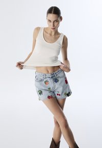 Top sin mangas blanco de canalé combinado con unos pantalones cortos de denim azul desgastados que presentan un bordado colorido de frutas y un ajuste relajado. Botines marrones hasta el tobillo.