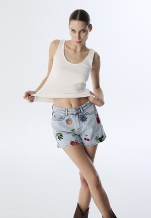 Un débardeur blanc à côtes associé à un short en denim bleu déchiré orné de broderies de fruits colorés et d'une coupe décontractée. Des bottes chelsea marron.