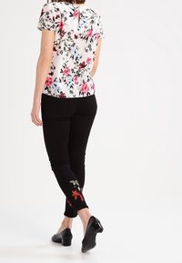 Floral blouse with short sleeves in cream, featuring pink and blue flowers. Black pants with red floral embroidery at the cuffs. Black heels.