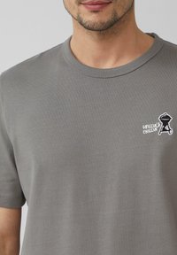 Szara bawełniana koszulka z okrągłym dekoltem. Posiada małe czarno-białe logo grilla z napisem "Grillin’ & Chillin’". Delikatna, ribbowana struktura.