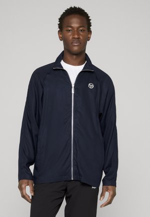 Mand iført en marineblå jakke med lynlås over en hvid t-shirt og sorte bukser, stående mod en ensartet grå baggrund.