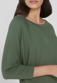 Olivegrünes Langarmoberteil mit rundem Halsausschnitt und weichem, glattem Stoff. Hat einen lockeren Schnitt und leicht hochgekrempelte Ärmel.