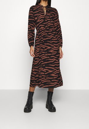 Kvinde iført en langærmet, sort og brun zebrastribet midi-kjole med ankelhøje, sorte snørestøvler, stående mod en ensfarvet baggrund.