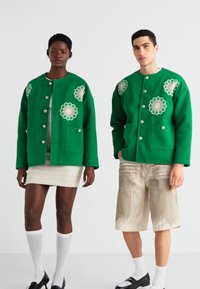 Grønn ullblandingsjakke med broderte blomstermønstre, oversized passform og sølvknapp-lukking, kombinert med et teksturert skjørt og lyse bukser.