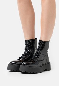 Bottes de combat en cuir verni noir avec une semelle noire épaisse, présentant un laçage à l'avant, un bout arrondi et un détail de fermeture éclair sur le côté.