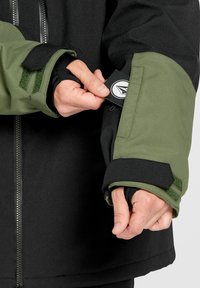 Giacca nera e verde con superficie texture, dotata di zip e chiusura con velcro sui polsini. Una mano tiene un'etichetta dalla tasca della giacca.