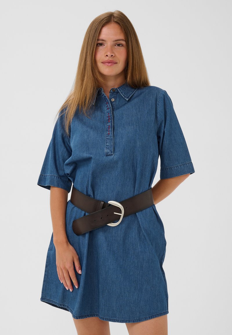 Frau mit glattem hellbraunem Haar, die ein blaues Denimkleid mit halblangen Ärmeln und einem breiten dunklen Gürtel mit silberner Schnalle trägt.