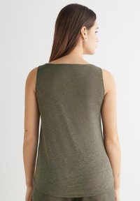 Olivově zelený bezrukávový top s kulatým výstřihem, měkkou texturou a volným střihem. Lem mírně zakulacený na zadní straně, s minimálním šitím.