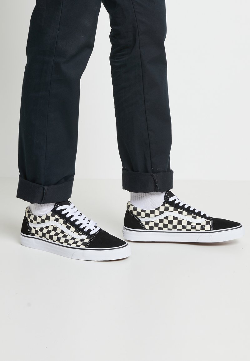 Personne portant un pantalon noir et des chaussettes blanches, avec des baskets basses à carreaux noir et blanc et des lacets blancs sur un sol blanc.