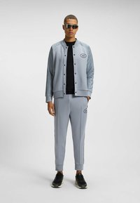 Grå sportjacka med rutiga ärmar, cirkulär logotyp. Matchande grå byxor med sidostråk, bärs med svarta sneakers.