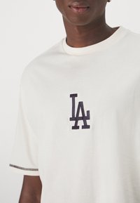 Bílý bavlněný tričko s námořnickou výšivkou loga "LA" na hrudi, s kontrastním prošíváním podél okrajů rukávů.