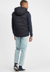 Schwarze Pufferweste mit Kapuze, aus gestepptem Material und geradem Schnitt. Kombiniert mit hellblauen Jeans und marineblauen High-Top-Sneakern.