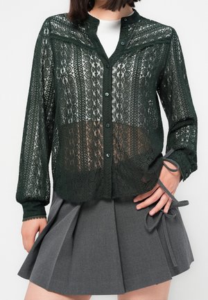 Femme portant un chemisier en dentelle vert foncé à boutons par-dessus un top blanc, associé à une jupe grise plissée avec un détail de nœud noué.