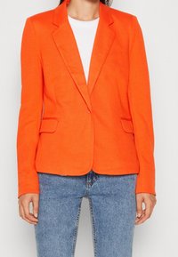 Blazer orange avec des revers structurés, deux poches avant et une coupe ajustée. Associé à une chemise blanche côtelée et un jean bleu clair.