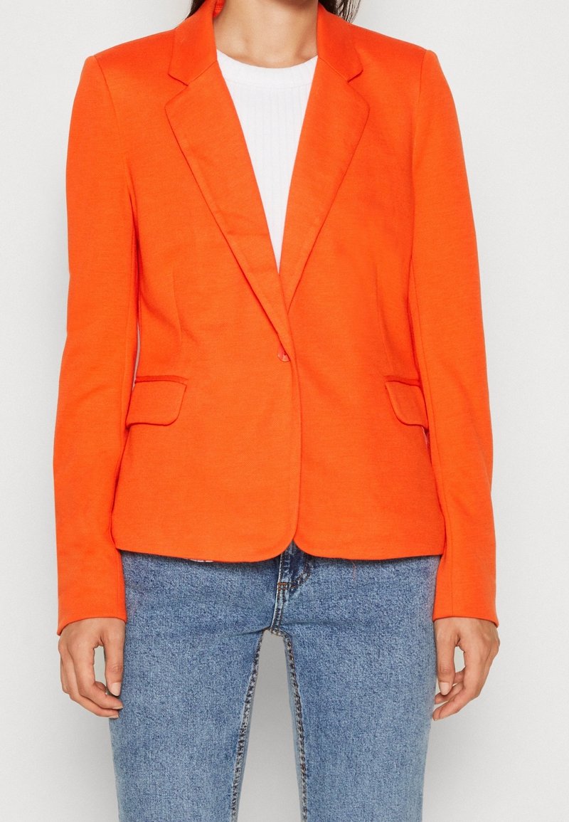 Blazer orange avec des revers structurés, deux poches avant et une coupe ajustée. Associé à une chemise blanche côtelée et un jean bleu clair.