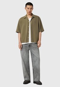 Khaki overhemd met korte mouwen en knoopsluiting, voorzien van een patch pocket, gecombineerd met losse grijze denim jeans en zwarte schoeisel. Effen achtergrond.