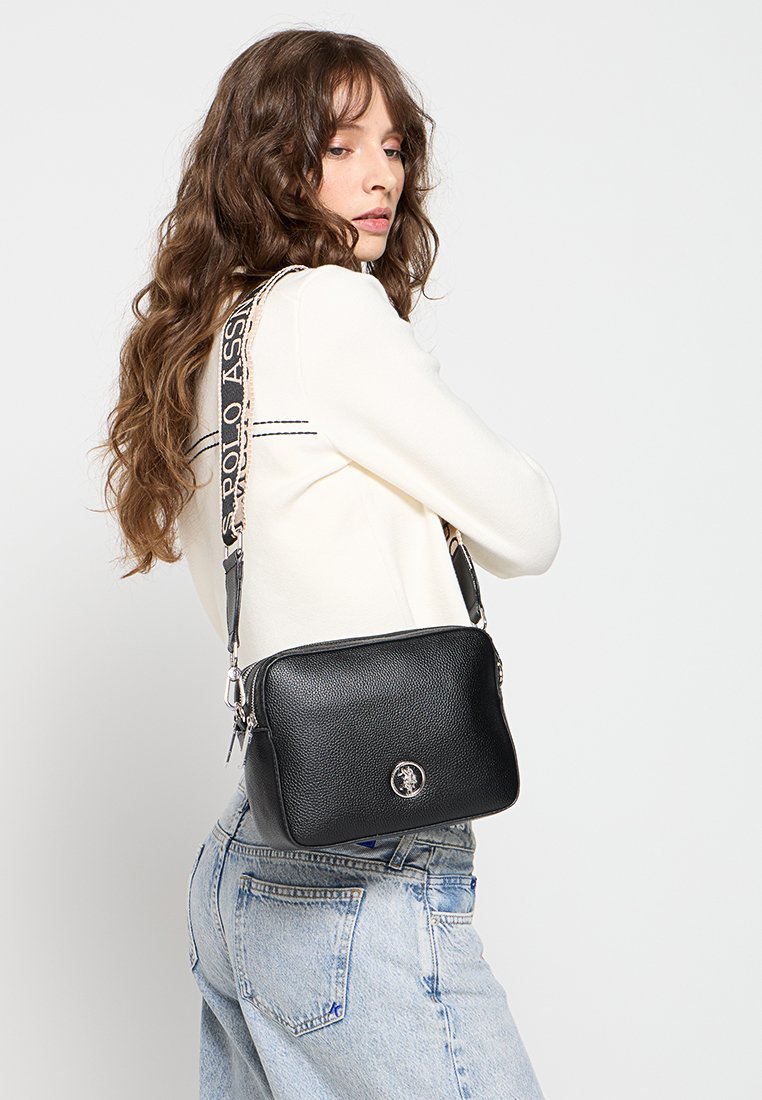 Bolso bandolera de cuero negro con un acabado texturizado, que presenta un acento de logo y una correa estampada. Forma rectangular compacta.
