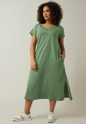 Kobieta o kręconych włosach, ubrana w zieloną midi sukienkę z krótkim rękawem oraz białe trampki, stojąca z jedną ręką w kieszeni, patrząca w lewo.