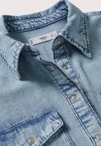 Chemise en denim bleu clair avec un col classique, des boutons-pression argentés et une poche. Présente une texture subtilement délavée et des détails de coutures.