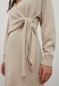 Robe en tricot beige à manches longues, avec un design croisé et un détail de nœud sur le côté. Sa texture douce et ses poignets côtelés complètent le look.