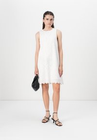 Robe blanche sans manches avec un motif ajouré, ourlet en scallop, associée à des sandales noires à lanières et un petit sac noir. Texture lisse.