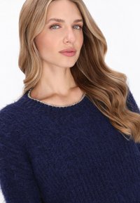 Marineblauwe, fuzzy gebreide trui met een gestructureerde afwerking en een ronde halslijn, versierd met zilveren accenten rond de kraag.