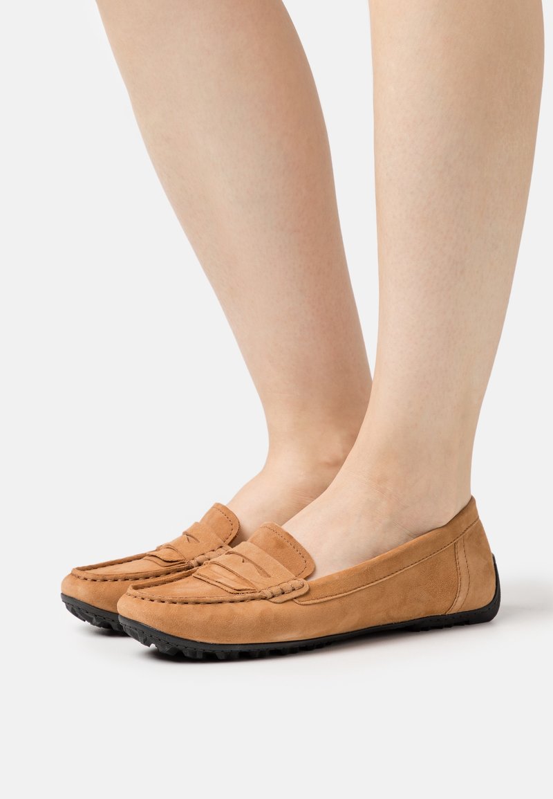 Mocassins en daim beige avec une semelle noire flexible, présentant un design cousu et une finition lisse, portés sur des jambes nues.