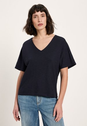 Frau mit kurzem dunklem Haar, die ein schwarzes V-Ausschnitt T-Shirt mit kurzen Ärmeln und blaue Jeans trägt, vor einem schlichten Hintergrund stehend.