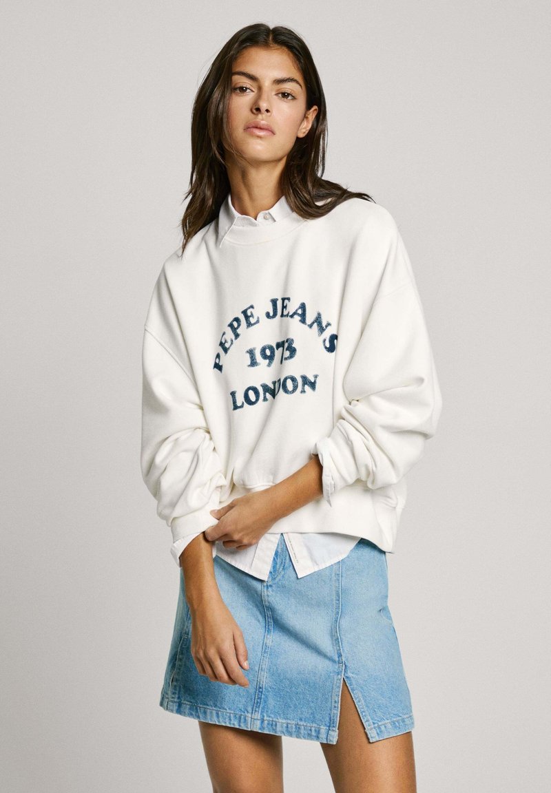 Sudadera blanca con texto bordado en azul "Pepe Jeans 1973 London", combinada con una falda de mezclilla azul claro con una abertura lateral. Corte casual.