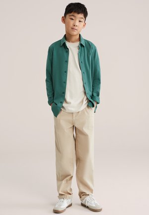 Zielona koszula na guziki na kremowej koszulce, do tego beżowe spodnie i białe trampki. Koszula ma lekko teksturowaną tkaninę.