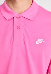 Rosa polo-skjorte lavet af struktureret stof. Har en klassisk krave, to rosa knapper og et hvidt Nike-logo på venstre bryst.