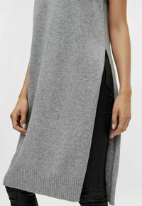 Grå stickad ärmlös tunika med ribbad nederkant, sidoflikar och textilstruktur, tillsammans med svarta åtsittande leggings.