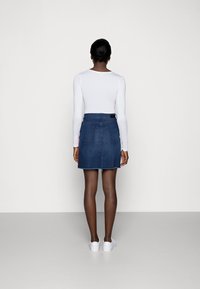 Džínová sukně s roztrhaným lemem, modré barvy, pěti kapsami a subtilní zadní štítkem s logem. Kombinovaná s bílým topem s dlouhým rukávem a bílými botami.