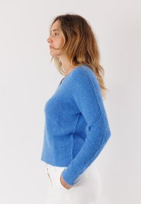 Maglione blu, morbido e a maglia, con scollo a V, maniche lunghe e vestibilità comoda. Indossato con pantaloni bianchi, che mettono in risalto il contrasto di texture e colore.