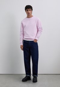 Hombre de pie contra una pared lisa, vestido con un sudadera rosa claro, jeans azul oscuro y zapatos de vestir negros, con una mano en el bolsillo.