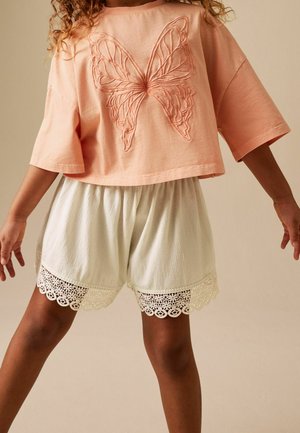 Gyerek, aki egy őszibarackszínű, túlméretezett inget visel pillangó hímzéssel és fehér texturált rövidnadrágot csipkés szegéllyel, sima háttér előtt áll.