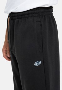 Czarne dresy wykonane z miękkiego materiału z elastycznym paskiem w talii, bocznymi kieszeniami i okrągłym logo po lewej stronie uda.