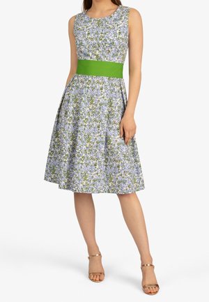 Kobieta w bezrękawowej, kwiatowej sukience do kolan z zielonym paskiem w talii i złotymi, otwartymi sandałami na obcasie, stojąca na białym tle.