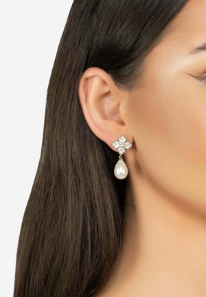 Orecchini a goccia dorati con un design floreale, arricchiti da cristalli trasparenti e una grande perla bianca nella parte inferiore, messi in risalto contro capelli scuri.