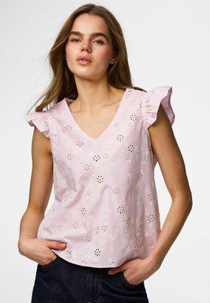 Hosszú barna hajú fiatal nő világos rózsaszín, ujjatlan blúzt visel virágmintás kivágásokkal és fodros ujjakkal, sötét farmerrel párosítva.