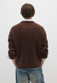 Brauner gerippter Strickpullover mit lockerer Passform und Kragen, kombiniert mit blauen Denim-Jeans mit einem ausgewaschenen Design und sichtbaren Gesäßtaschen.