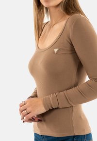 Camiseta de manga larga marrón de canalé con un escote redondeado y un pequeño logotipo triangular en el pecho. Presenta un diseño ajustado y una textura elástica.